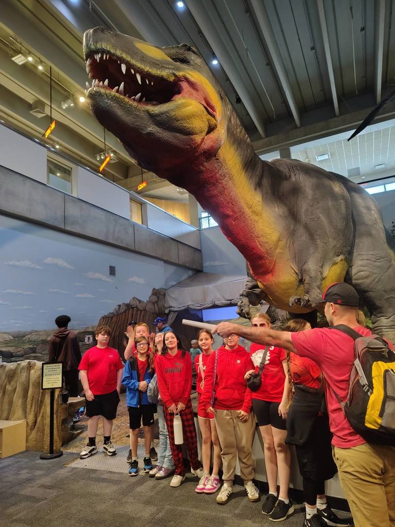 Several children in red jackets gather around an adult in a red shirt. They stand before a large dinosaur replica in a museum.