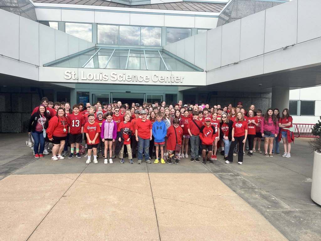 Large group wearing red shirts in front of a building. Some individuals have numbers on their shirts.