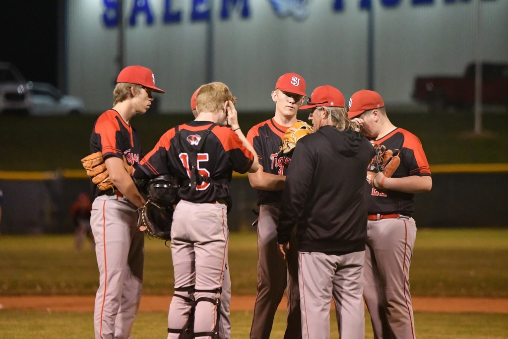 Baseball players in jerseys and caps gather near a field. A coach talks to them. Bright stadium lights illuminate the scene.
