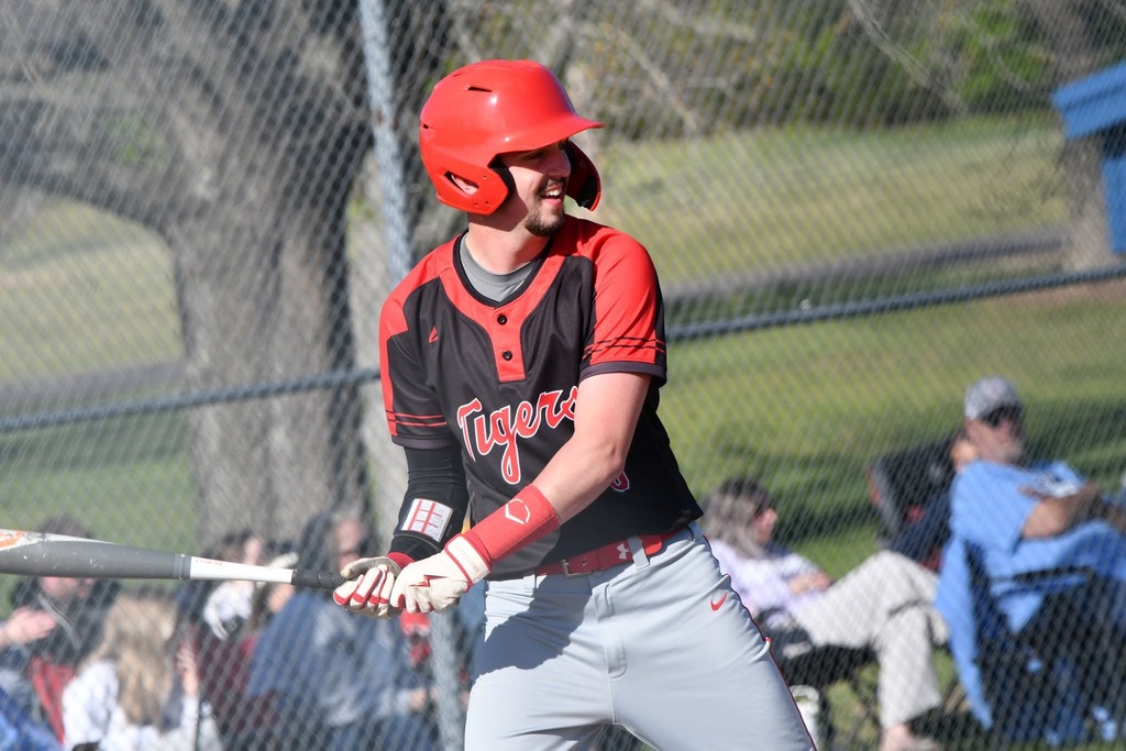 A baseball player in a red helmet and uniform swings a bat at a game. Spectators watch from behind a fence.