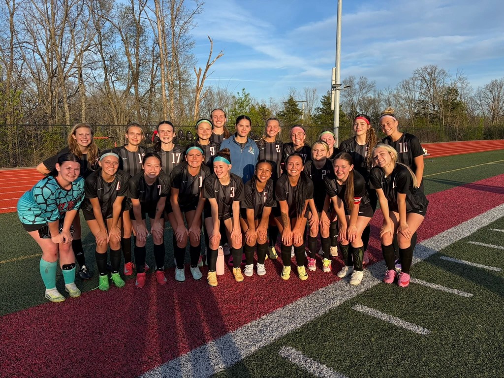 A soccer team in black uniforms stands on a field, with trees in the background.