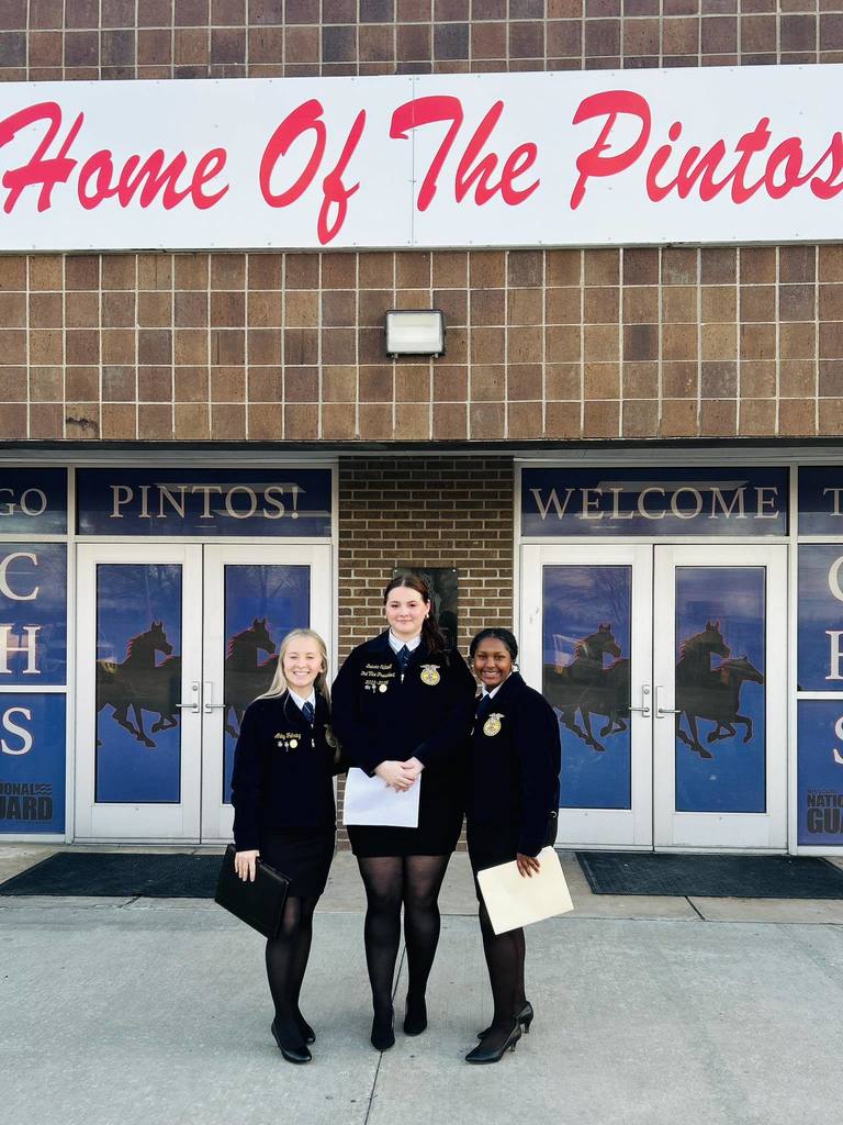 Three women in uniforms stand in front of a building with "Home Of The Pintos" sign.