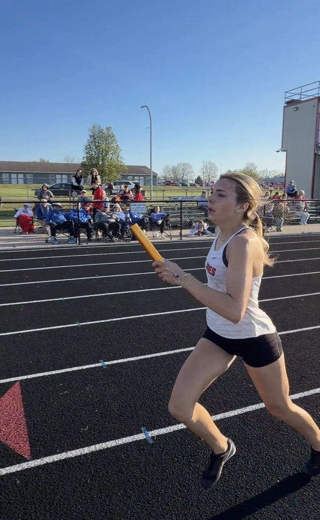 A female athlete runs on a track, holding a baton. Behind her, people sit on bleachers.