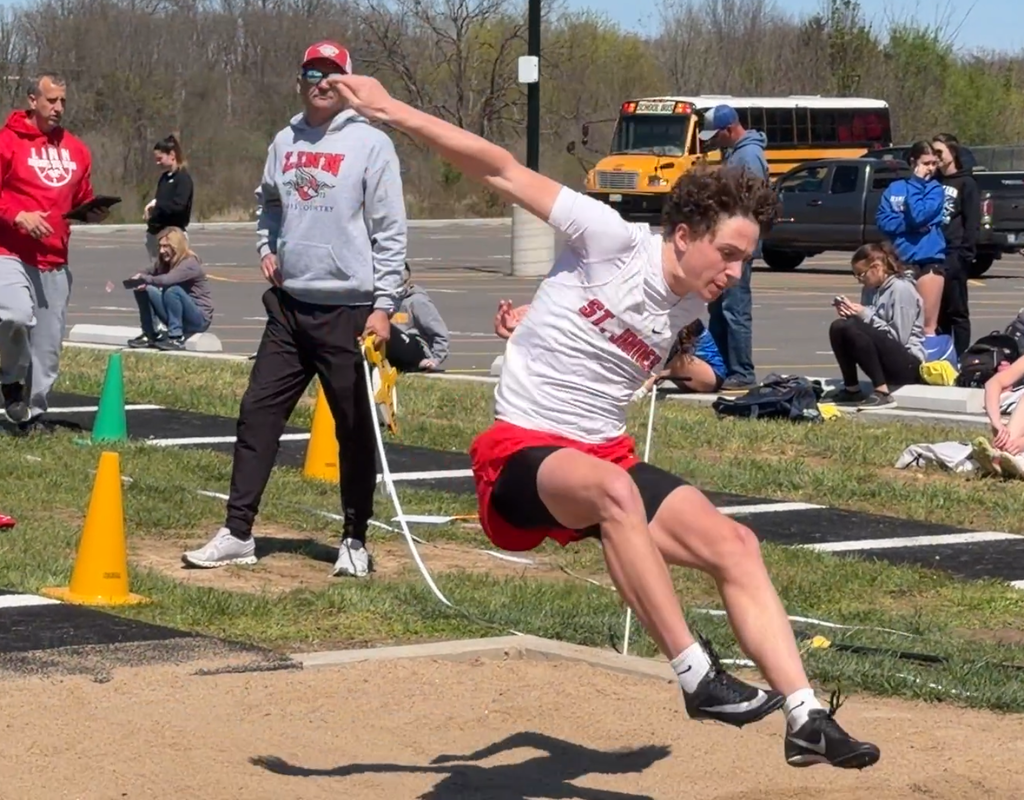 An athlete jumps in a sand pit wearing a white shirt, red shorts, and black sneakers.