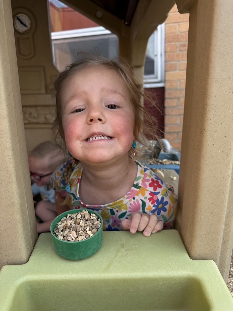 A girl smiles inside a playhouse, with a bowl of food in front. Behind her, a baby sits in a stroller.