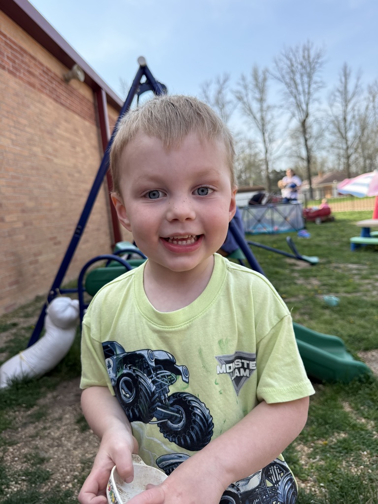 A smiling child in a yellow shirt with a monster truck on it stands in a playground with a building behind.