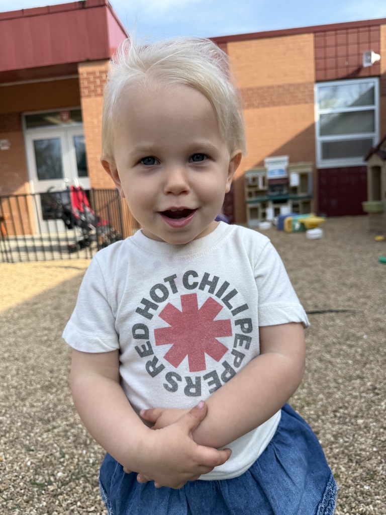 A toddler wearing a white shirt with a band logo stands in front of a building.