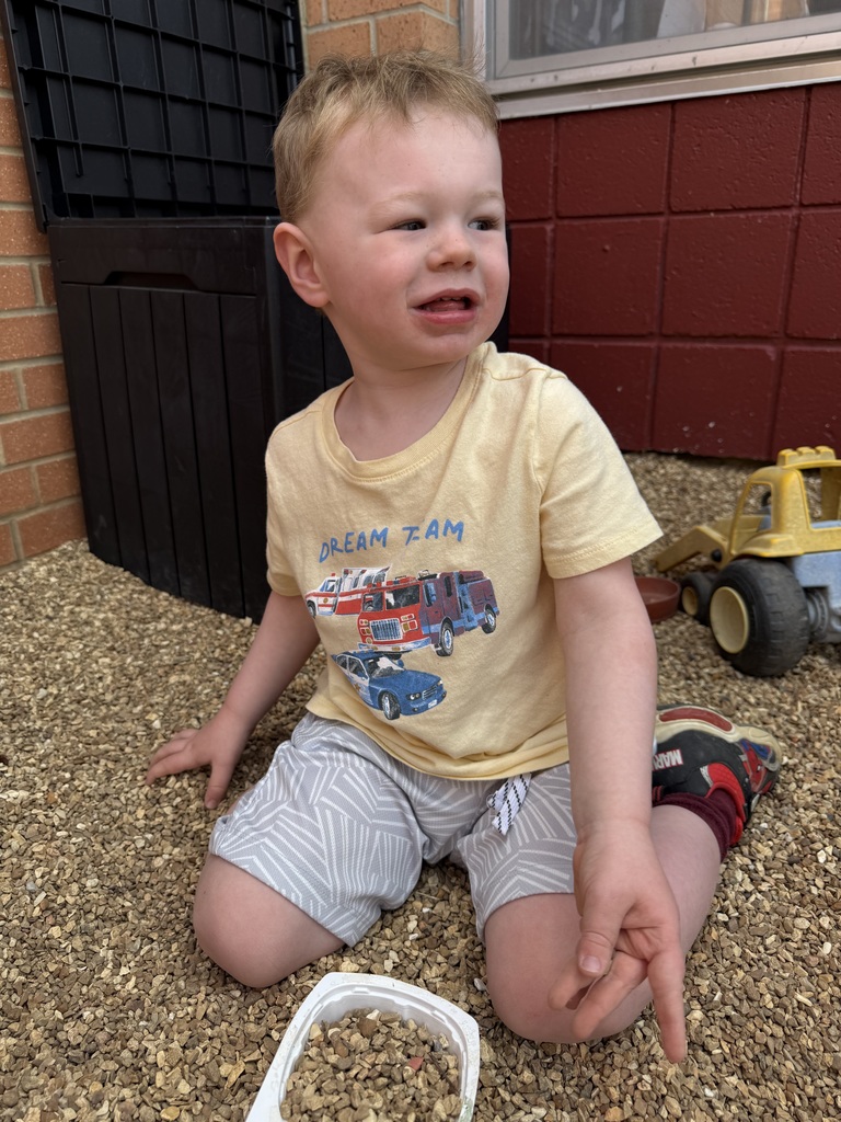 A child in a yellow shirt sits on gravel near toys and a brick wall.