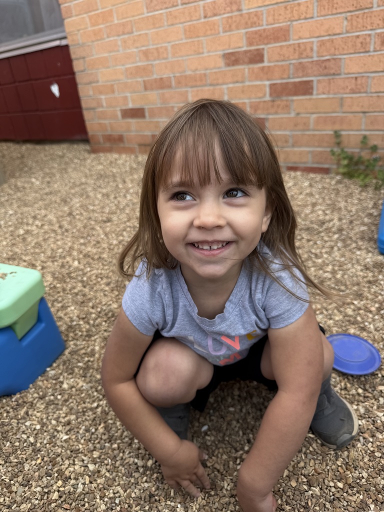 A young girl kneeling on the ground in front of a brick building with a big smile.