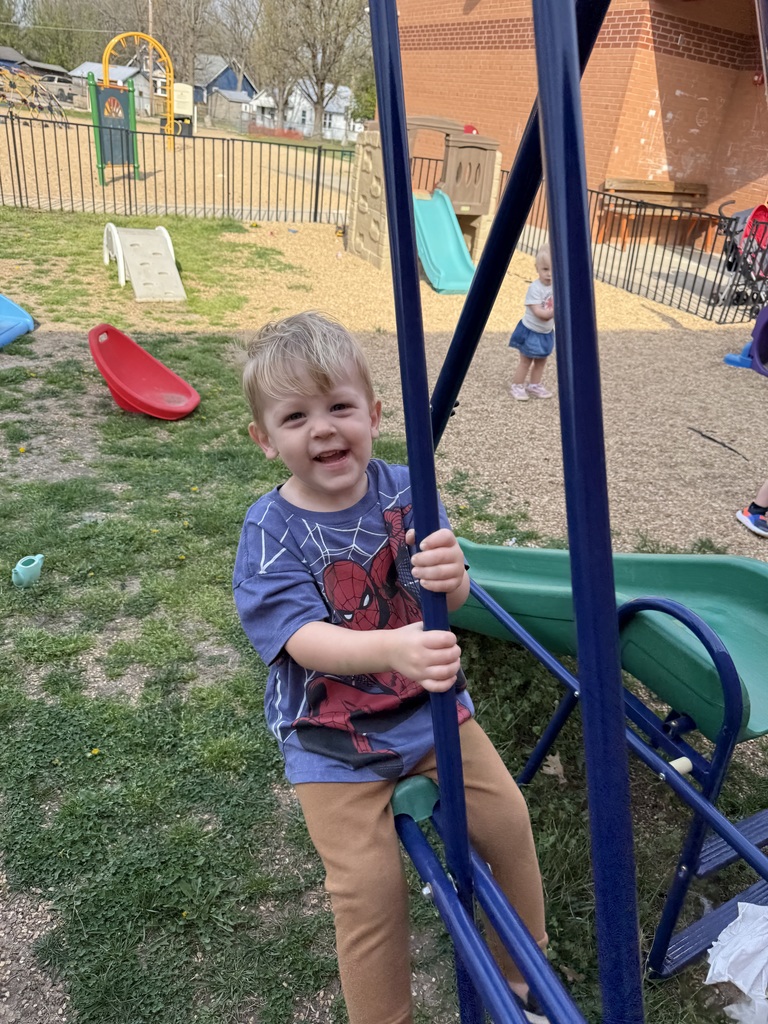 A young child with brown hair stands holding onto a blue pole at a playground. Behind them, another child plays on a green slide.