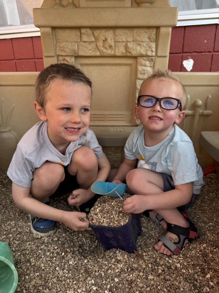 Two boys are sitting on the ground, playing with a small bowl of seeds. One wears glasses. Behind them is a stone wall.