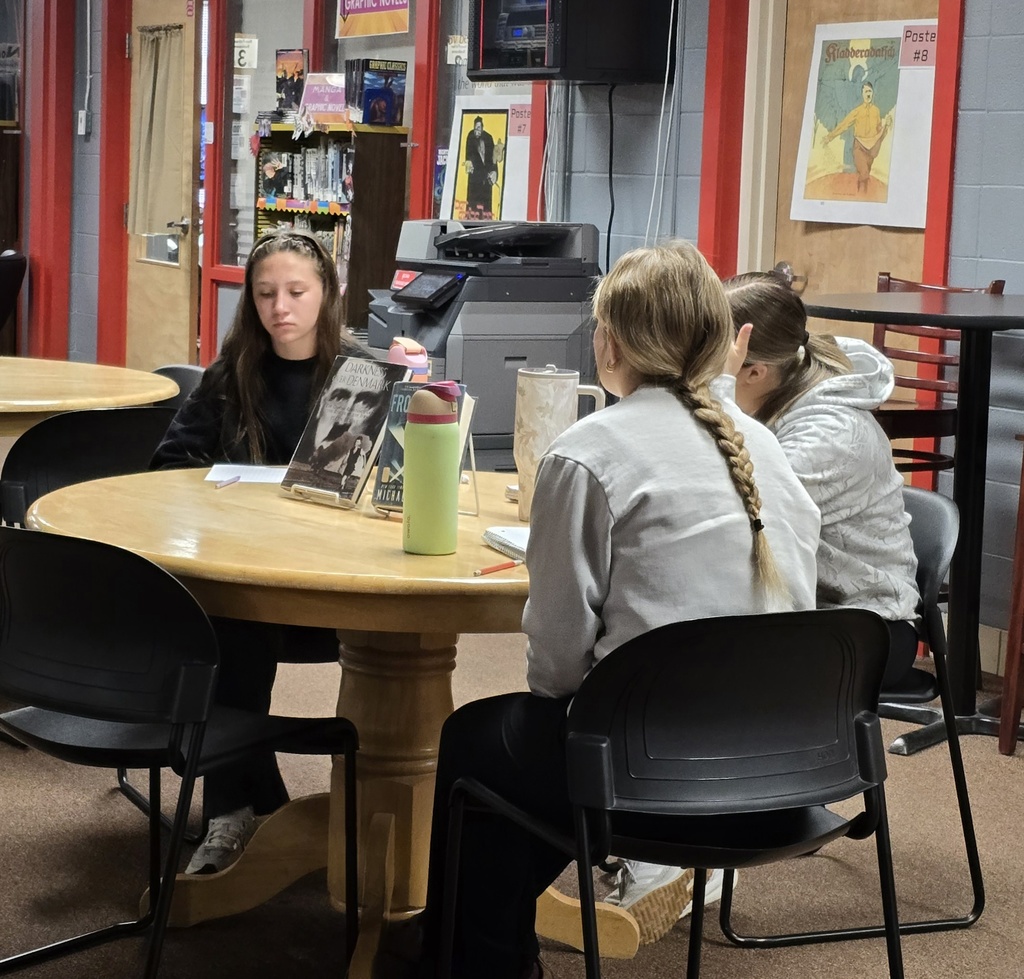 Three young people sit around a wooden table in a room. A woman sits with a book, a pen, and a cup.