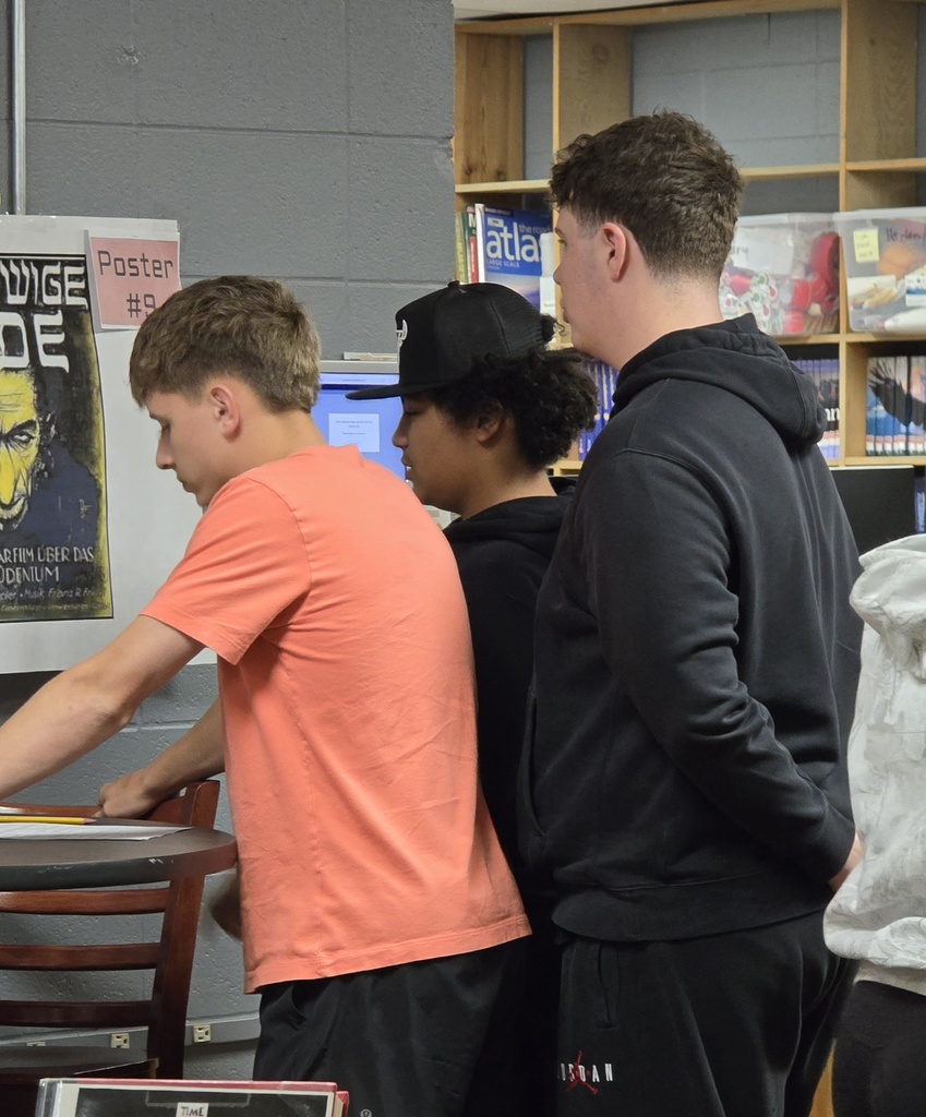 Three individuals near a shelf; one with an orange shirt, another in black with a cap, and a third in black with a hood.