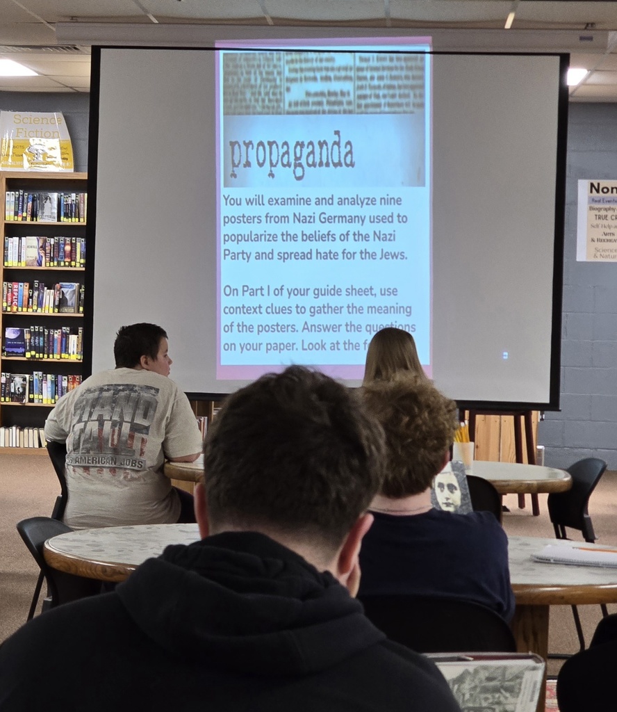 A classroom with students seated around tables. A large screen shows text related to Nazi propaganda. Shelves with books in the background.