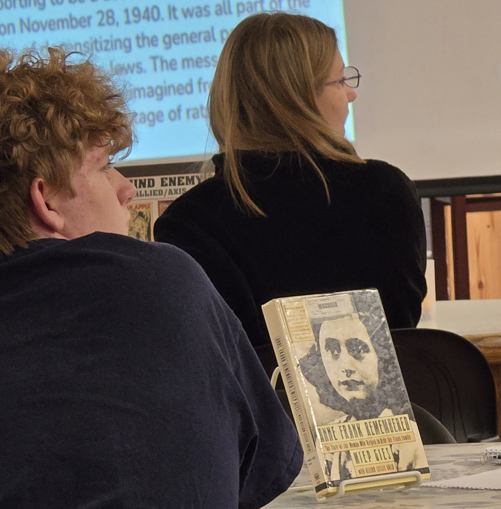 Two people in a classroom, a man with curly hair, and a woman with glasses. A book is on the desk.