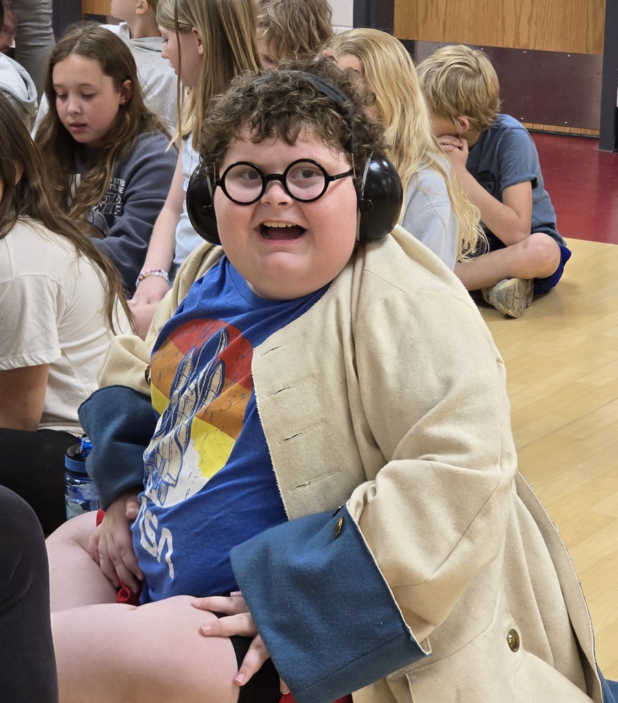 A person in a tan coat sits on a gym floor with friends. They wear glasses and headphones.