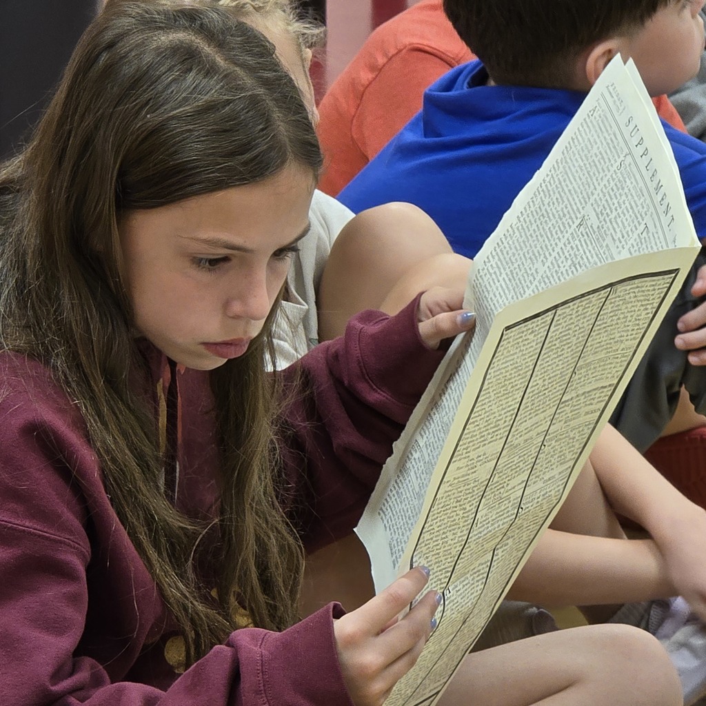 A girl reads from a book while seated in a classroom; others sit behind her.