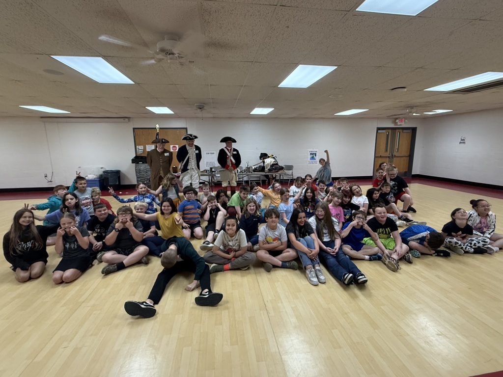 Many children seated in a large room with wooden floors. A few adults stand behind them. Bright lighting illuminates the space.