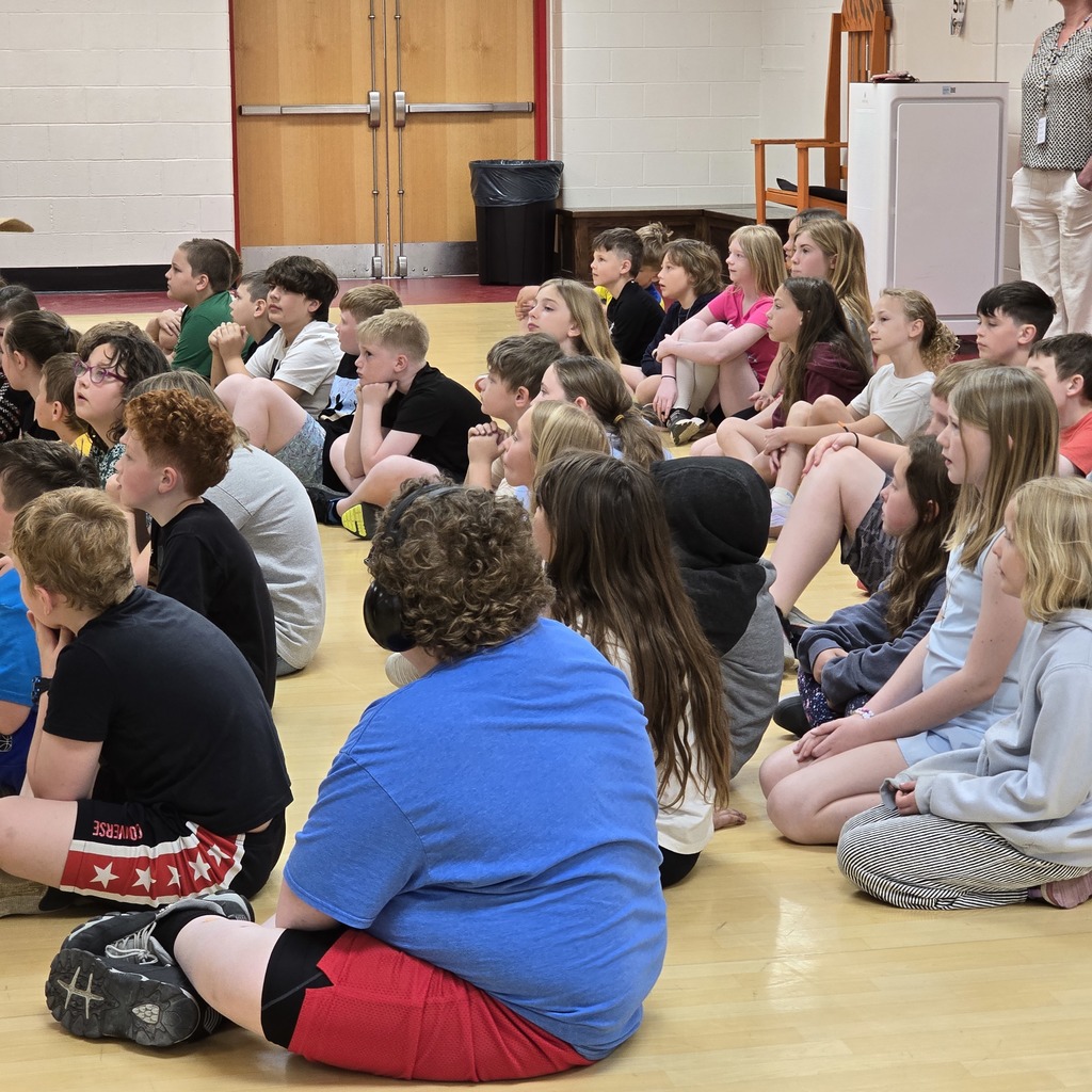 Many children sit on a floor in a room with wooden doors and a woman in the background.