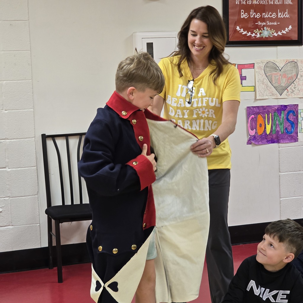 A young boy in costume stands with a woman, who holds a white cloth. Behind them, a boy sits, observing. Background includes a wall with framed art and a chair.