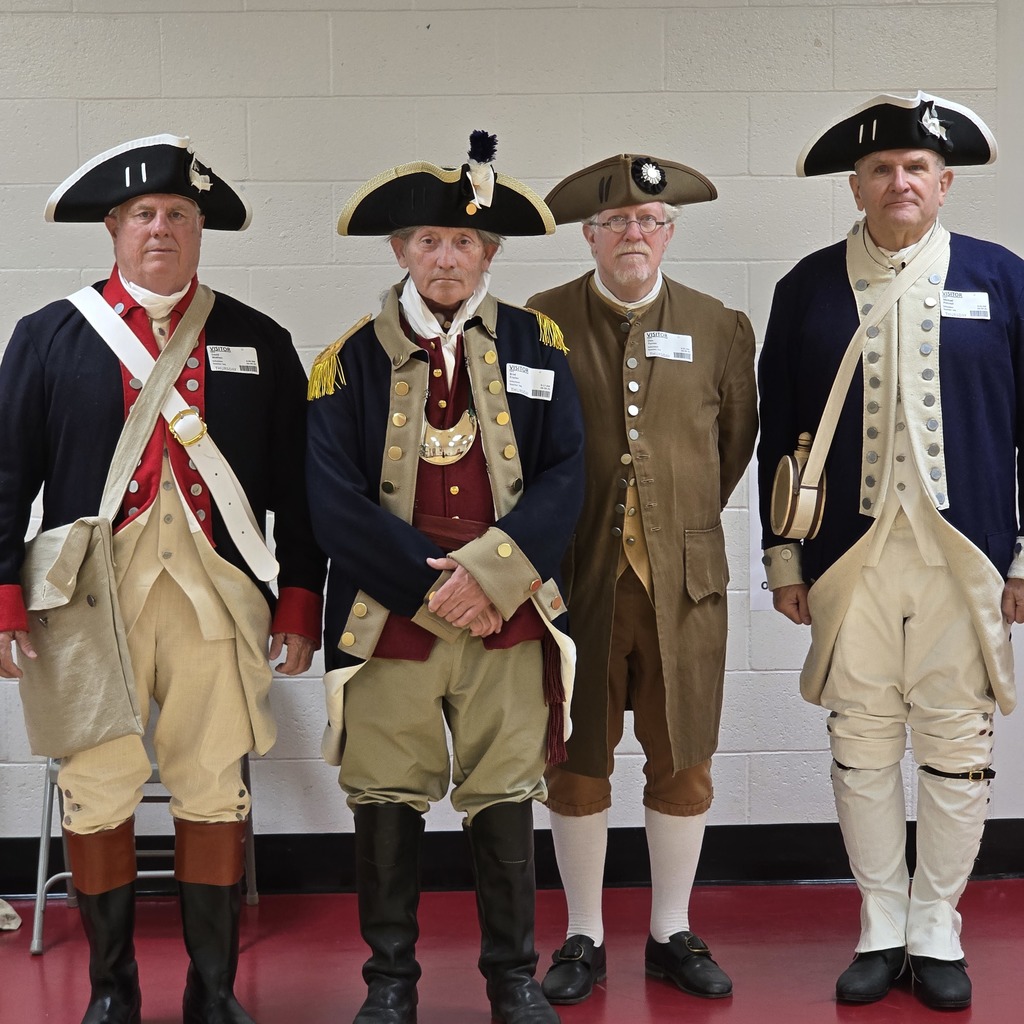 Four men in historical reenactment costumes pose together indoors. They wear hats and shoes, with uniforms varying in color and design.