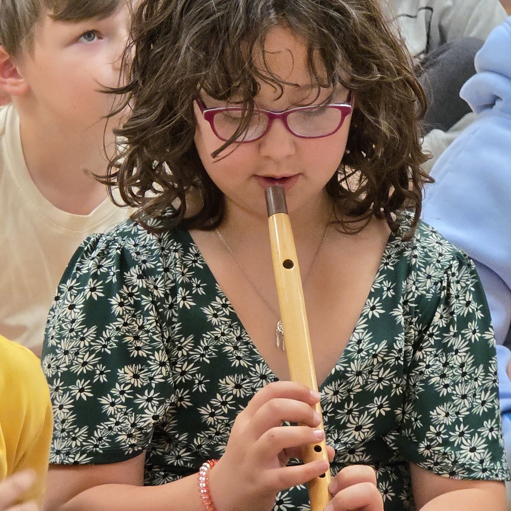 A girl in a floral-printed green shirt plays a flute while others look on. Background includes two other children.
