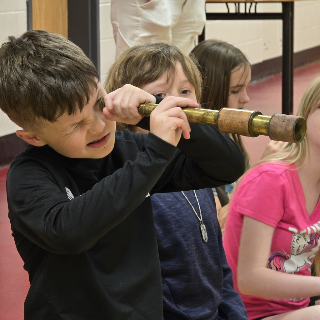 Child using a telescope inside classroom, other kids sitting behind. Bright red floor.