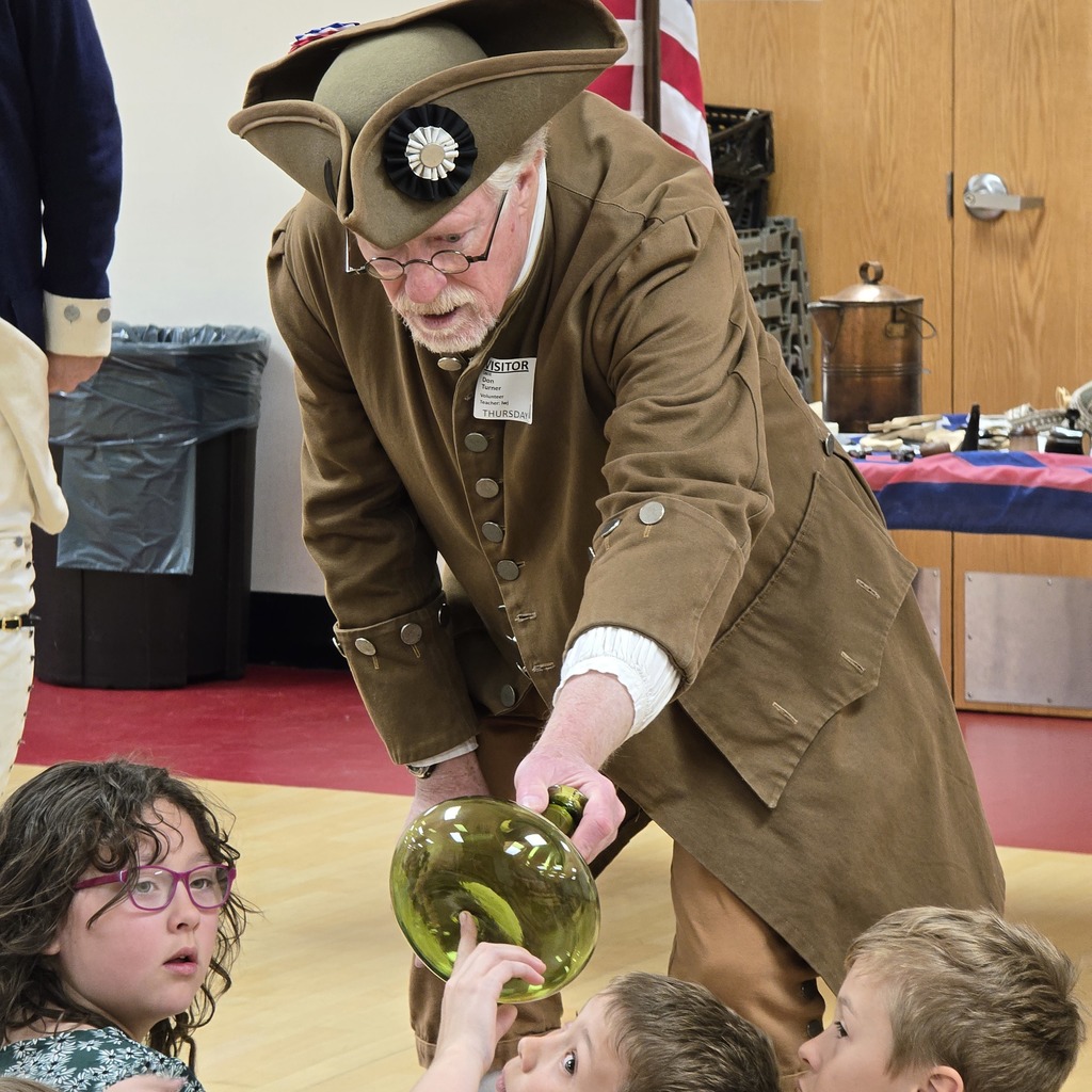 Person in costume showing a glass jar to children. Background includes a wooden wall, a flag, and a trash bin.