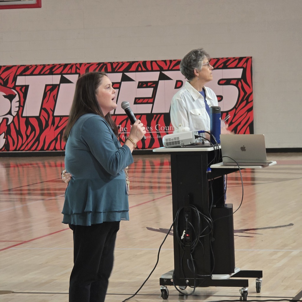 Woman speaking into a microphone, another woman standing behind a podium with a laptop in a gym.