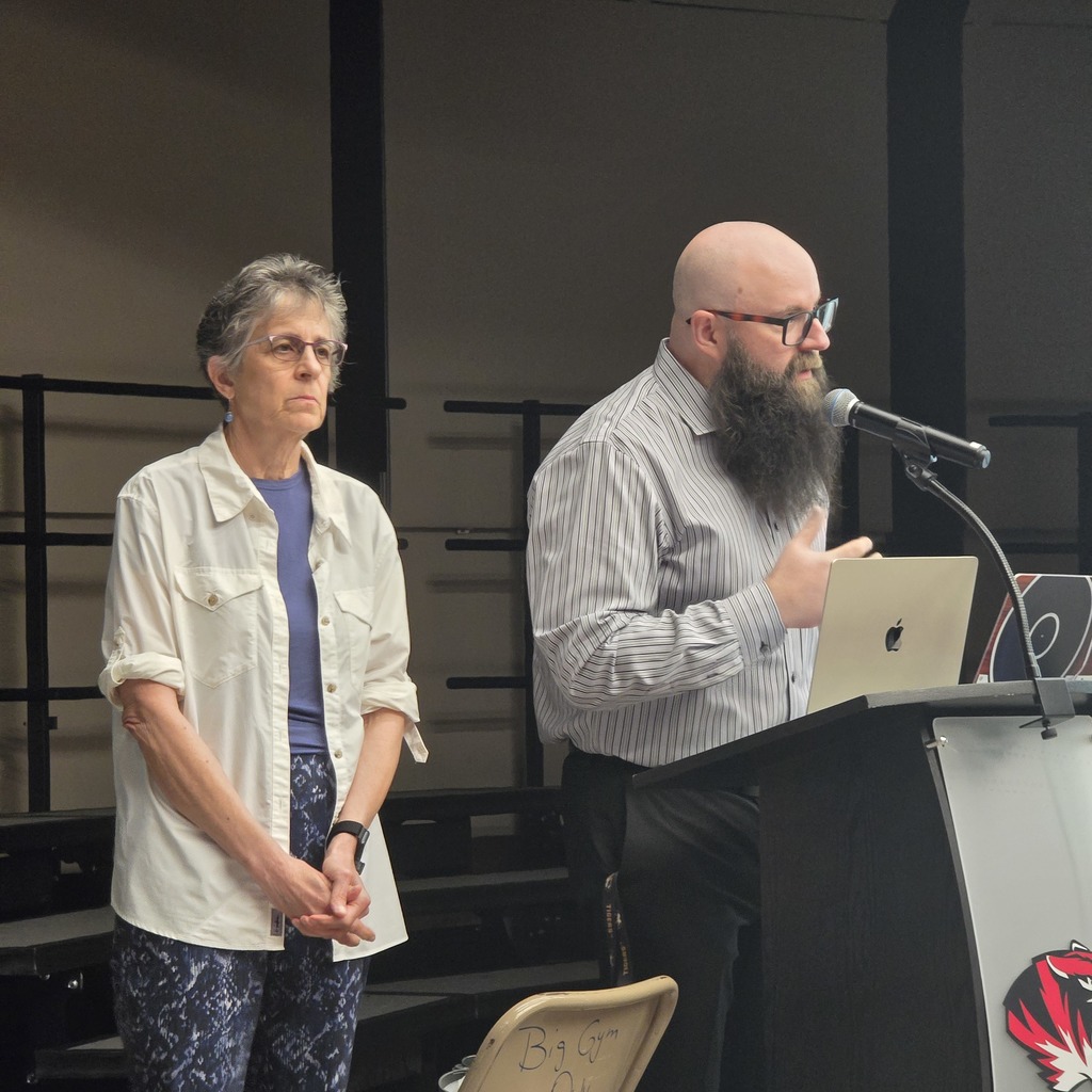Two people on stage with a podium. One person holds a laptop. The other person stands with a microphone.