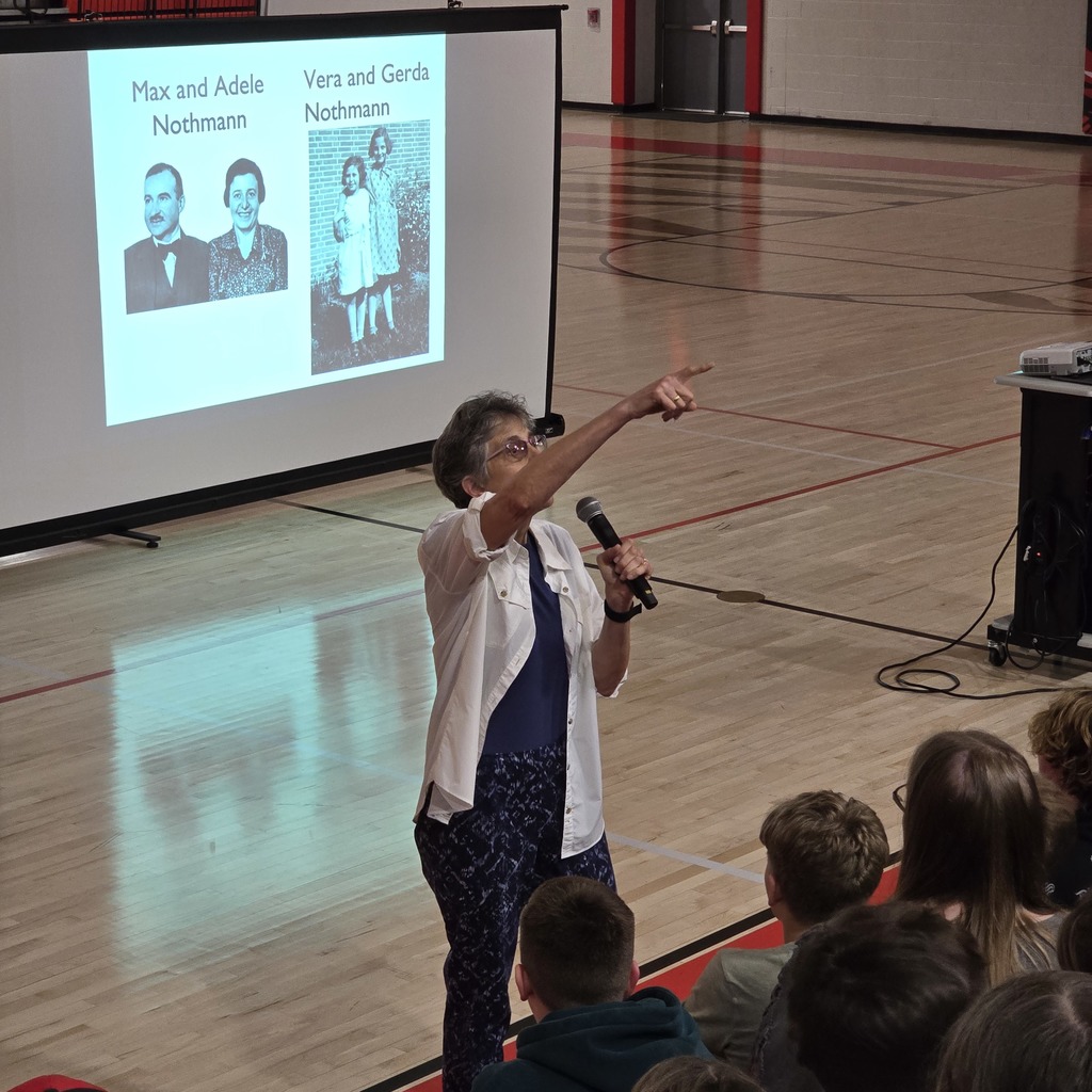 Woman speaks into a microphone in a gym with a projector screen showing pictures of two families.