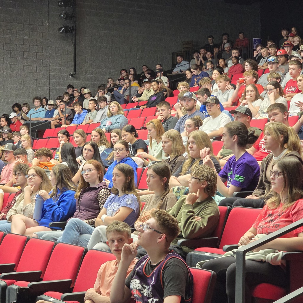 Auditorium filled with people seated on red chairs. Most wear casual clothing. Some wear glasses.