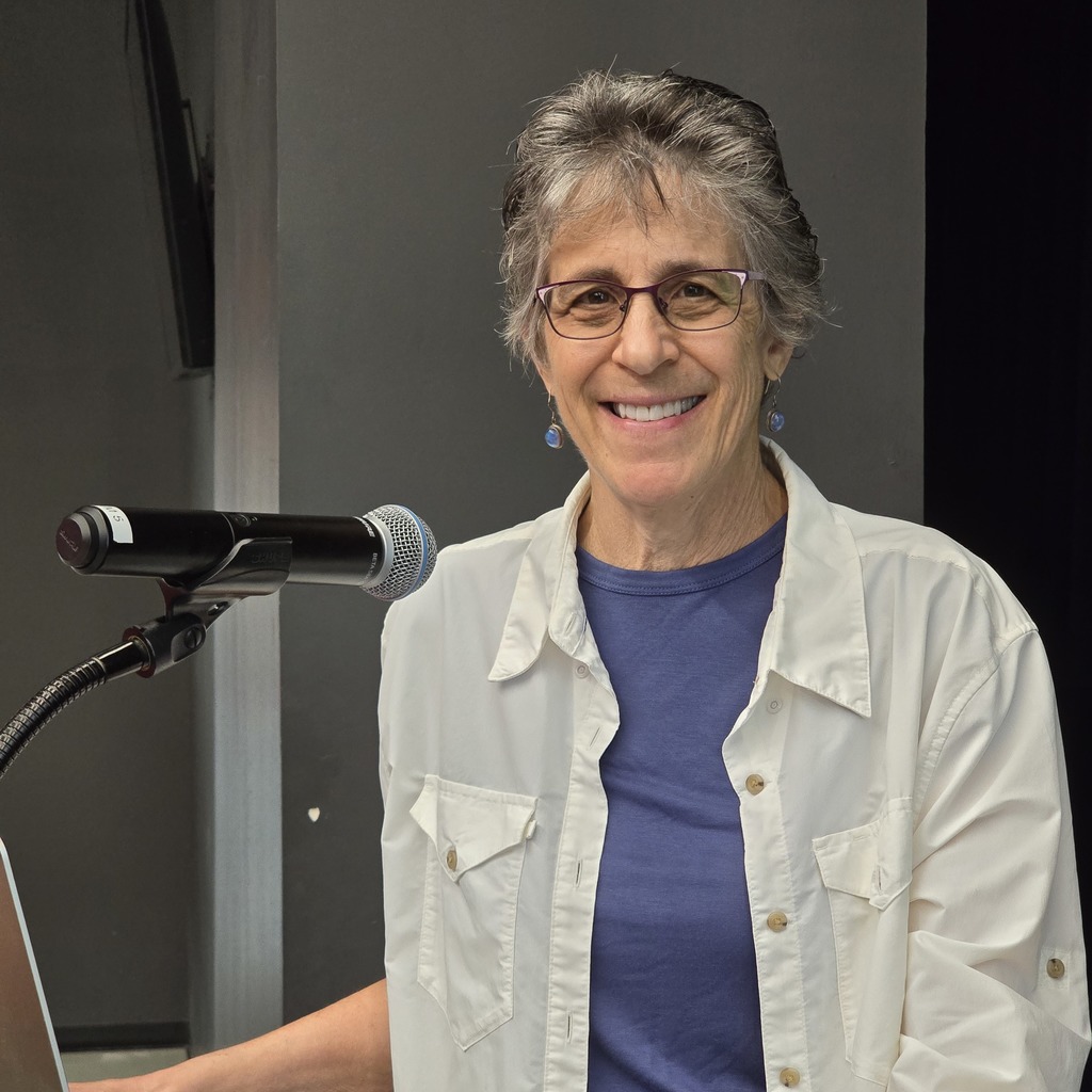 Woman wearing glasses and a white jacket standing at a podium with a microphone.
