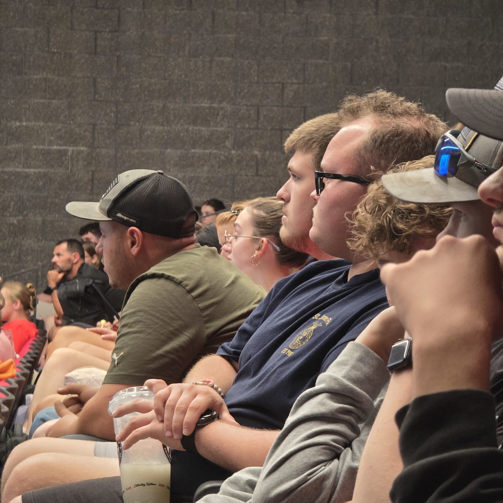 Group of people sitting in theater seats; some wear hats and glasses; one holds a beverage.