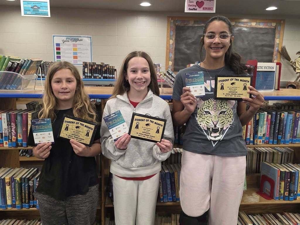 Four girls in a library, each holding a certificate, smiling. Shelves filled with books in the background.