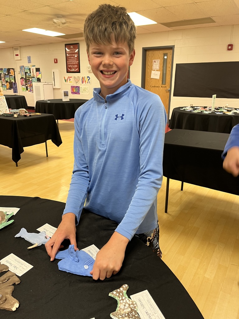 A child wearing a blue long-sleeve shirt is smiling and standing behind a table with paper cutouts.