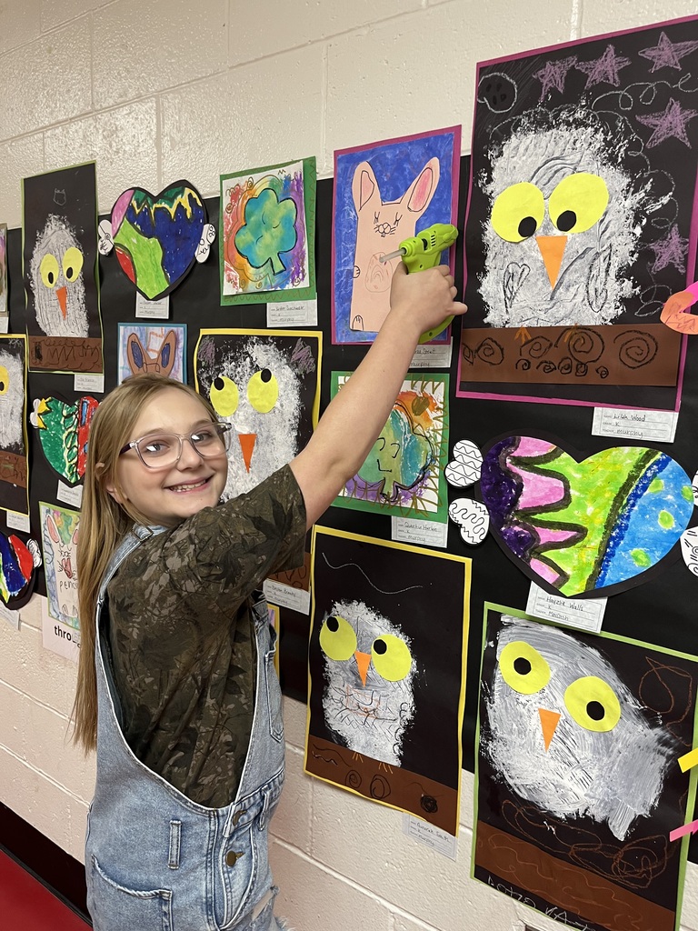 A young girl in glasses and overalls points to a colorful owl art on a classroom wall.