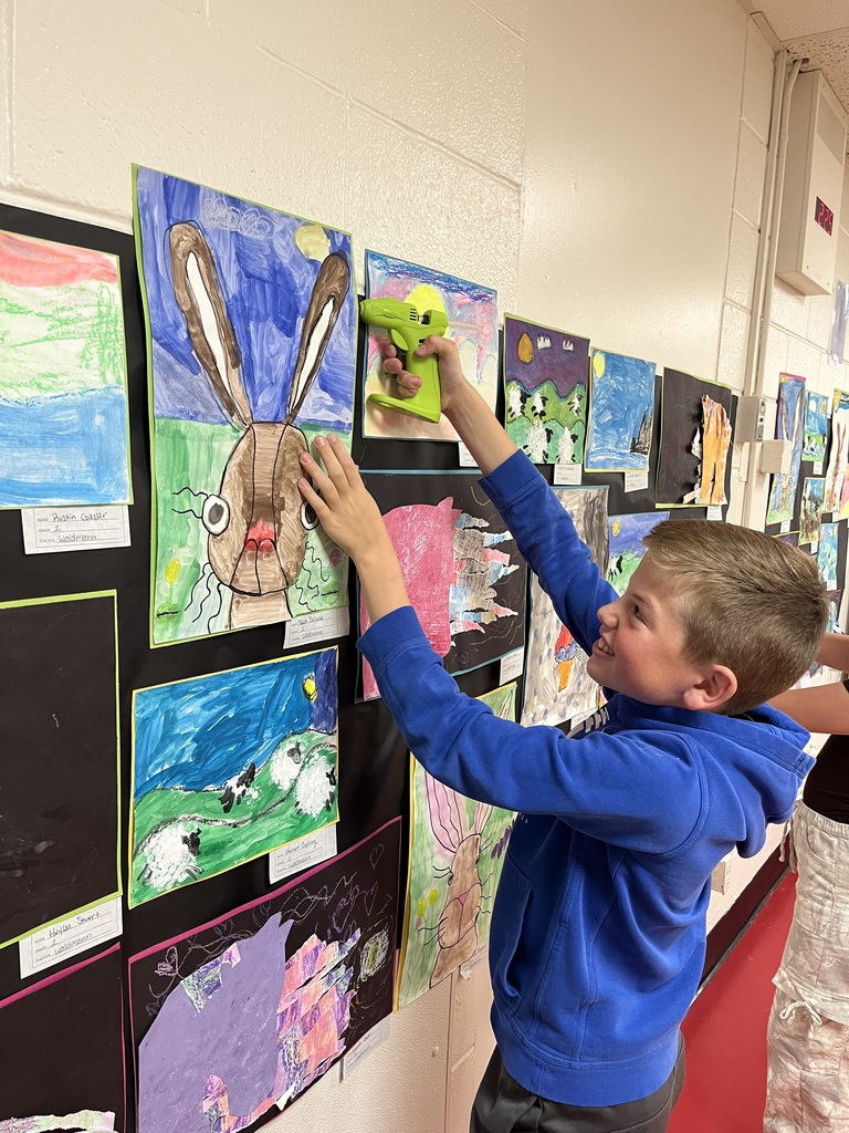 A young boy in a blue hoodie is pinning up a green bunny toy on a wall with several artworks.