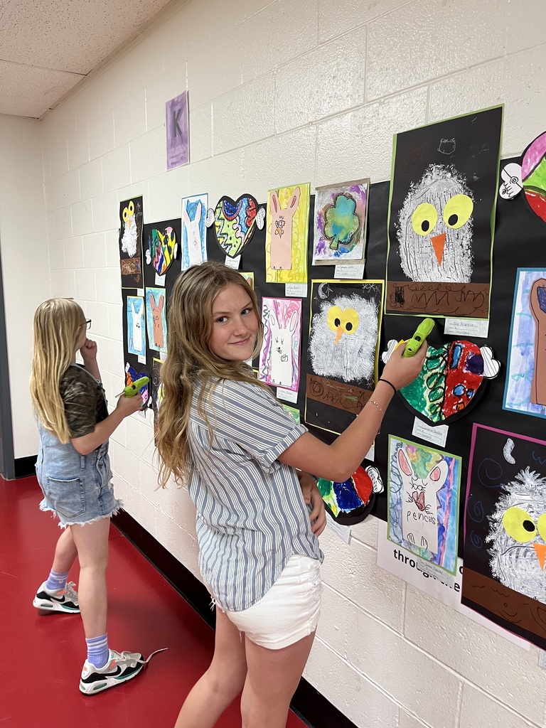 Two girls, one holding a glue stick, stand before a wall with artwork. One wears sneakers, the other denim shorts.