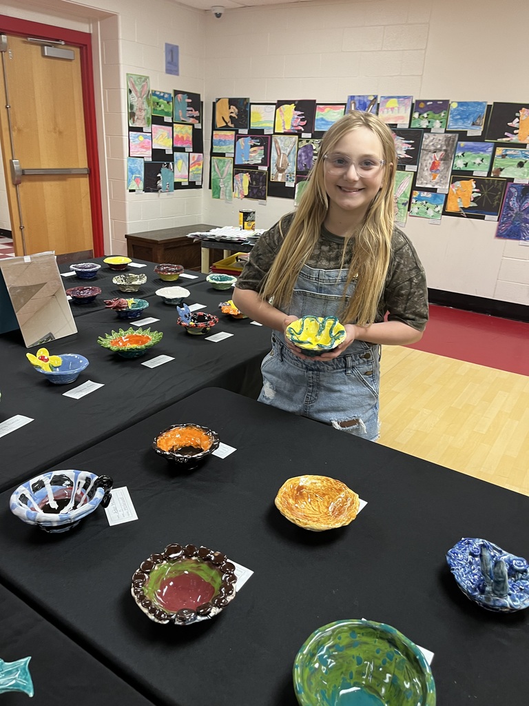 A young girl with glasses stands by a table displaying colorful ceramic bowls. The room has art on the walls.