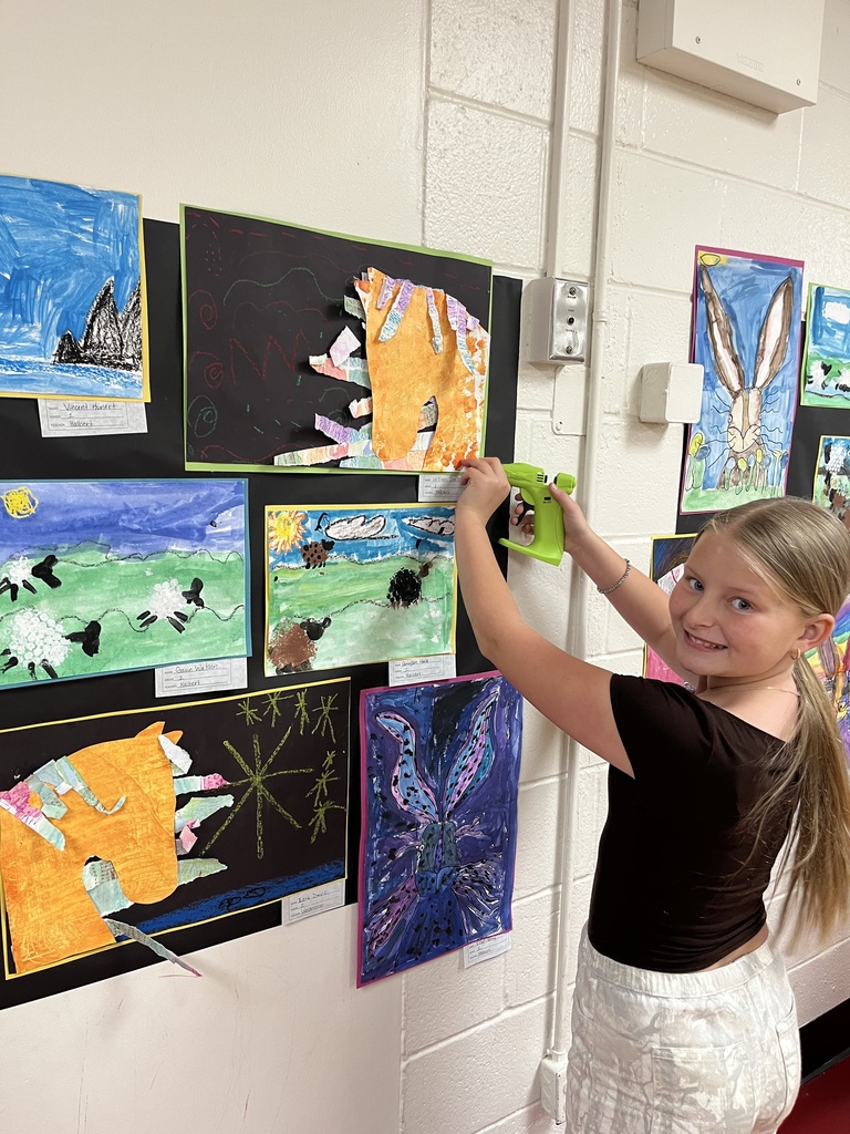 Girl in black top and white skirt pins artwork to wall. Many different pieces of artwork.