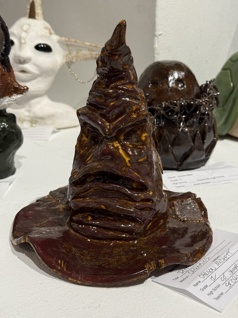 Ceramic pieces, including a hat shaped like a wizard's hat, sit on a table. A white bust and other pieces are behind the hat.