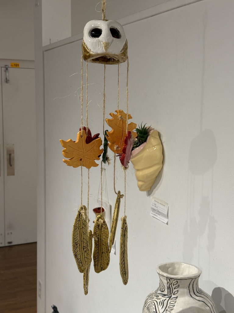 A wind chime with an owl face and leaves hangs on a wall, next to a small plant and a white vase.