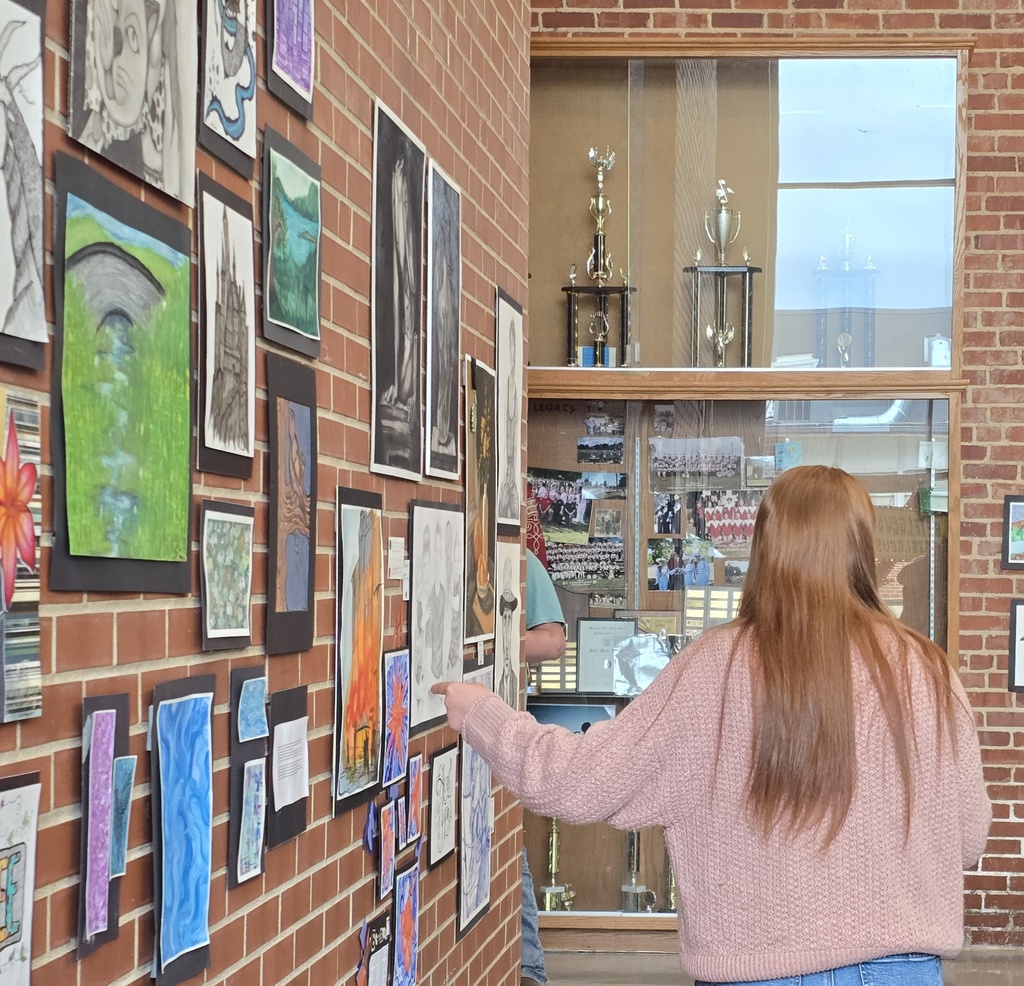A woman in a pink sweater points at a wall with framed artwork. A brick wall and glass display case with trophies in the background.
