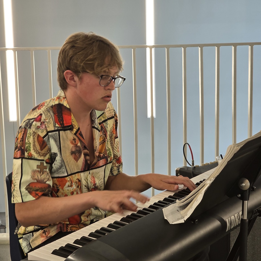 A person wearing glasses and a patterned shirt seated, playing a piano with an open music sheet.