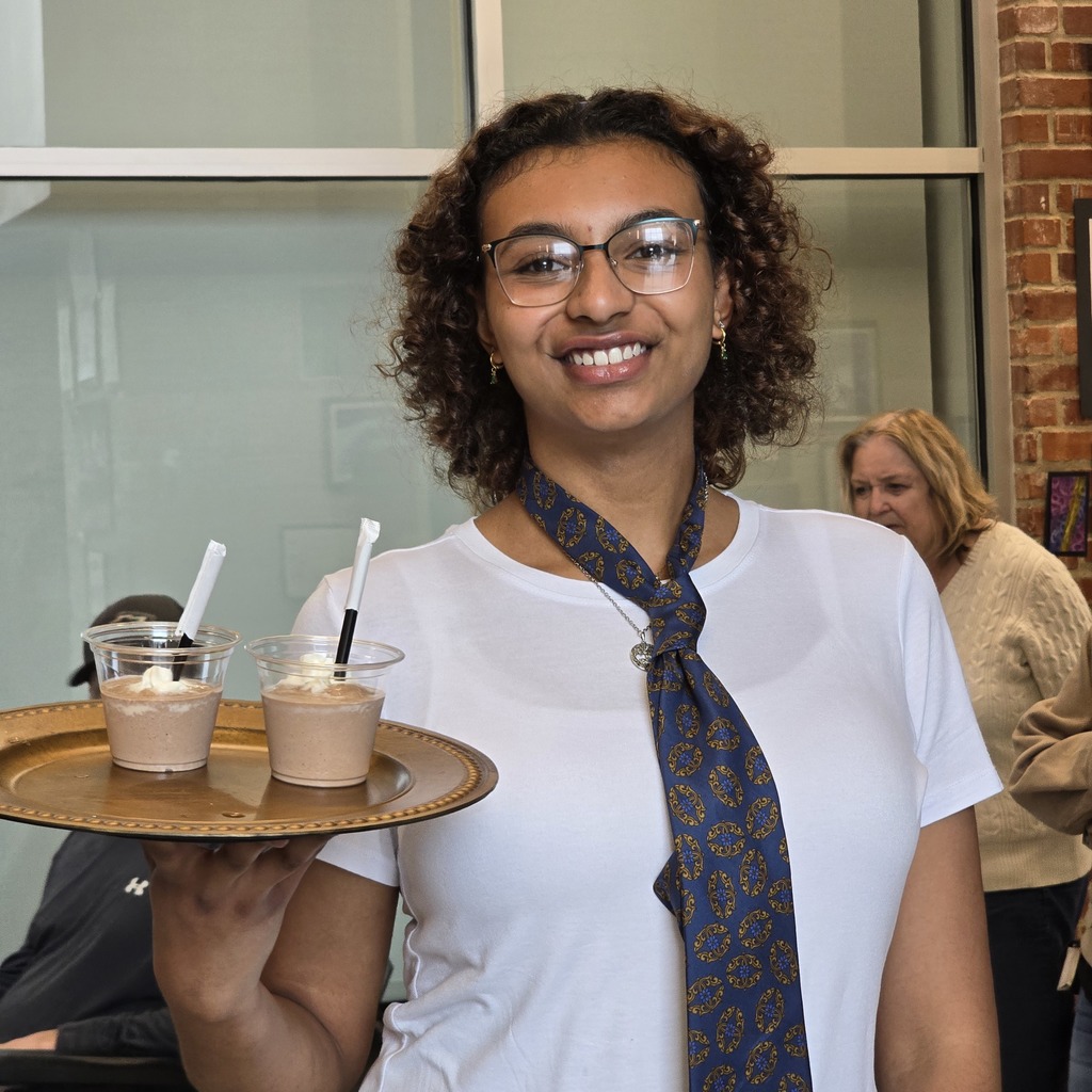 A person with curly hair holds a tray with three cups of dessert. They wear glasses and a blue tie.
