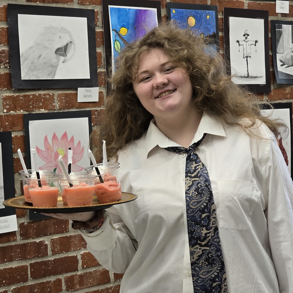 Woman with long curly hair holds a tray with small cups of pink drink in front of a brick wall with art.
