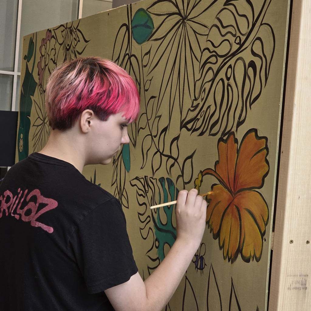 A person with pink hair paints a large wall mural featuring flowers and leaves.