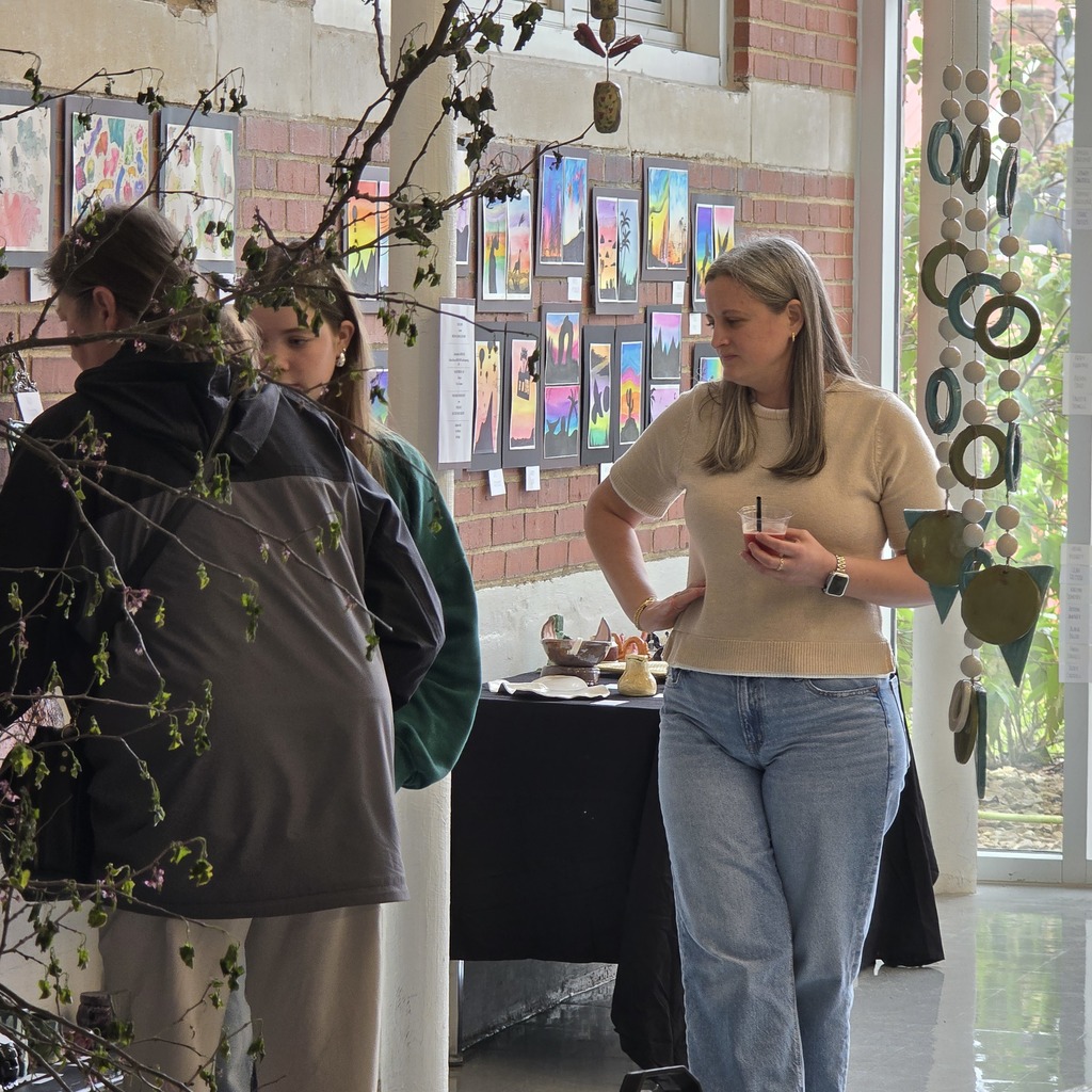 Three people view a wall with framed art. A woman in jeans holds a drink.