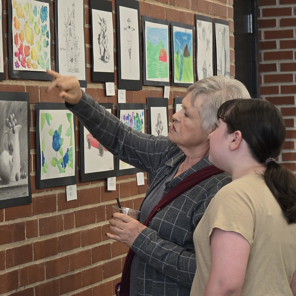 A woman with gray hair points to artwork on a brick wall while another woman with a ponytail watches.